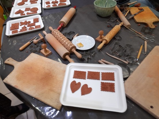 A creative chaos of dough rollers, some with Christmas decor, of dough stamps and tools for cutting and carving. Brown clay decorations wait on white cardboard plates for drying.