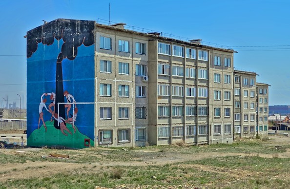 A five storey brezhnevka style housing block. The building has seen better days and the surrounding land is poorly maintained. On the side of the building is a painted mural of people dancing round an industrial chimney pouring black smoke in to the sky.