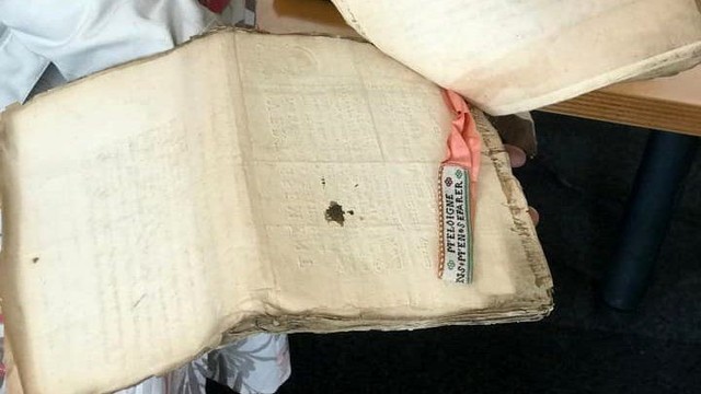 Une liasse de papiers liés dans l'angle supérieur gauche, tenue ouverte par une archiviste. Entre 2 papiers, une jarretière brodée, bordée en haut de fil rouge et en bas de fil vert, accrochée à la liasse par un ruban rose.
On lit une partie du texte :
"m'éloigne
ns m'en séparer".