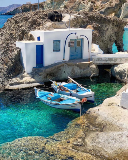 a photo of two small rowboats tied up on the shore, there is a building built into the rockface, the boats are white with some blue in them, matching the colors of the building, the water is aqua turquoise colored , the rocks light brown