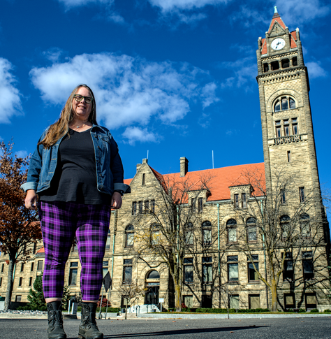 A woman in purple plaid pants and a jean jacket stands in-front of a building with a large clock tower.