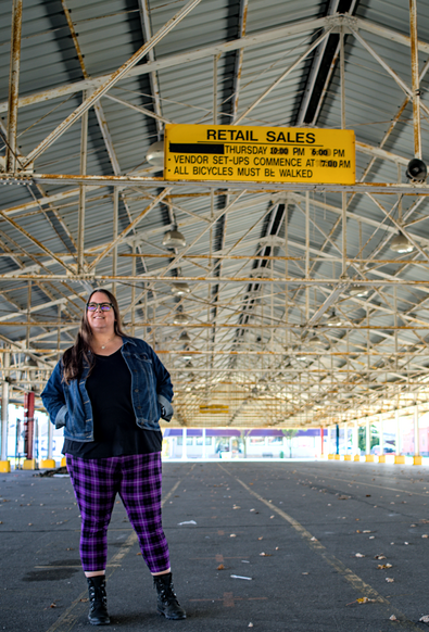 A woman in purple plaid pants and a jean jacket stands under a long metal awning.