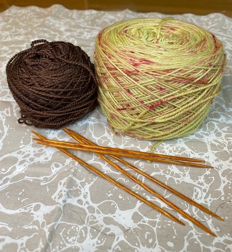 A full skein of wound yarn in yellow & reddish brown next to a mini skein of wound yarn in brown. 5 wooden double-pointed needles are in front