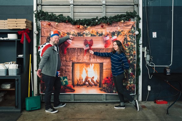 A newly minted American stands next to a fake fireplace decorated for Christmas next to his wife holding a miniature flag