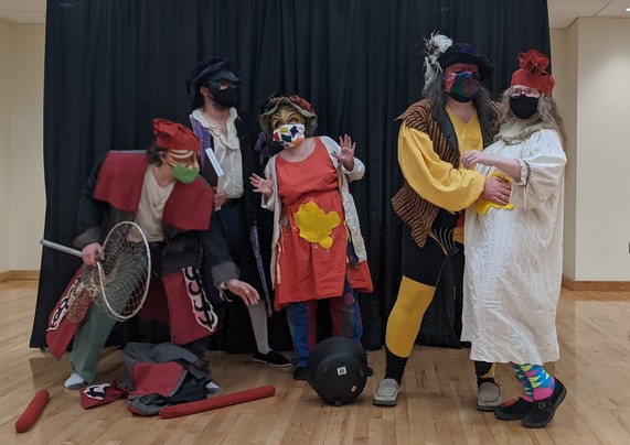 Five actors perform a commedia dell'arte play in front of a black backdrop in a university dance studio during a medieval history conference portraying the stock characters of Brighella (disguised as a warrior made of sausage), il Dottore, and Arlecchino (smeared with mustard) looking shocked as il Capitano smears mustard on Vittoria (in a chemise and partially disguised as the queen of sausage people).