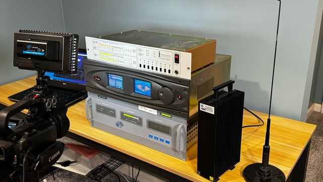 Video camera pointed at Harris Digit CD FM exciter, Omnia 6 broadcast audio processor, and Inovonics FM modulation monitor stacked on top of each other in operation on a table. Beside the stack is a Bird Termaline dummy load to prevent the RF from radiating. An antenna is placed next to the dummy load, connected to an SDR out of frame.