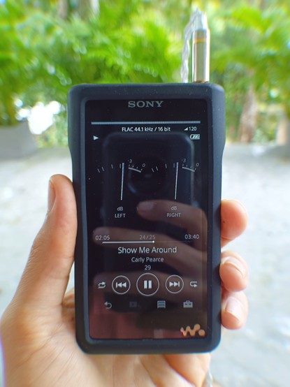 SONY walkman in my hand playing "Show Me Around" by Carly Pearce, with a view of our balcony and some trees and bushes in the backdrop.