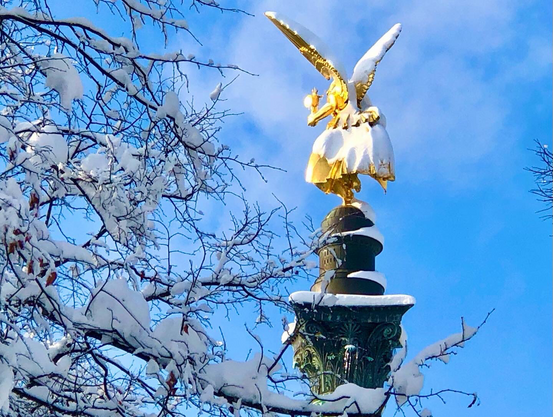 The gold statue of the Angel of Peace, her gown draped with snow, with trees in the background, all covered in snow, at Munich’s Friedensengel