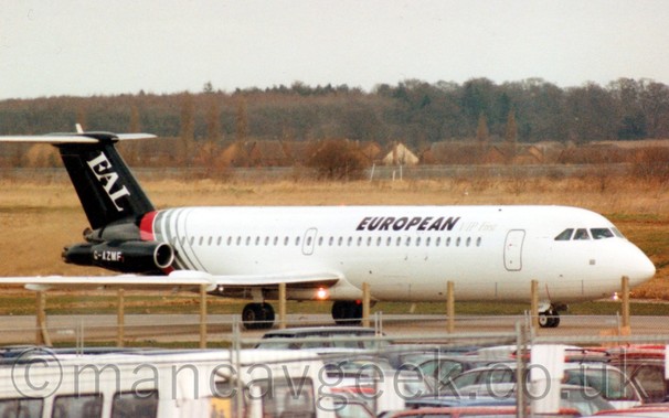 Side view of a white and black, twin engined jet airliner, with the engines mounted on the rear fuselage, taxiing from left to right, with a ground level car park in the foreground, , and yellowing grass fields running off to trees in the distance, under a hazy grey sky.