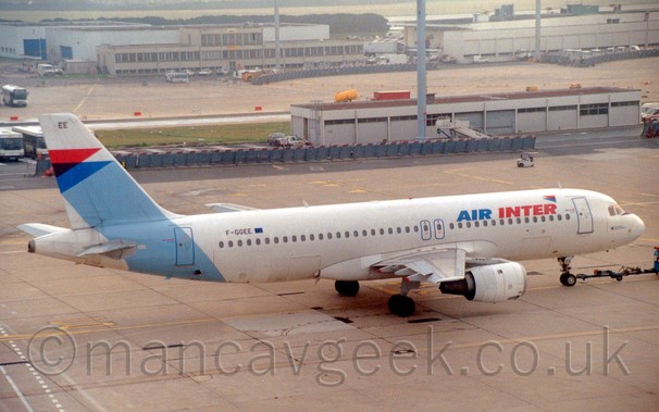 Side view of a white and blue, twin engined jet airliner being pushed back from it's stand by a truck by a pole attached to the nosewheel. with 2 and 3 storey buildings in the background slowly dissapearing into mist.