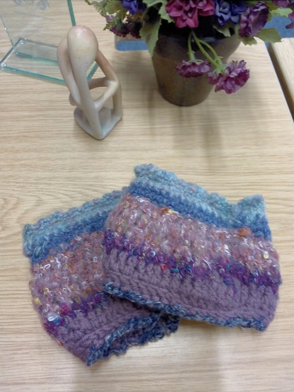 Crocheted cuffs (wristwarmers) in multiple colours of blue and pink. On a desk with a planter and a figure of a thinker and a glass award in the background.