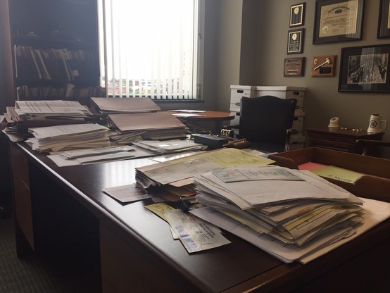 Color image of an office desk covered with stacks of documents. Center of the image is a brown wood executive desk with its surface stacked with documents. To the right in the image is a wall space covered with certificates, awards, and accomplishment plaques, in the center background is a window with blinds open showing an elevated cityscape and a wood bookshelf filled with files.