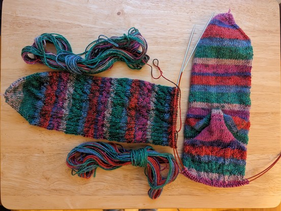 Two skeins of yarn with two socks being knit toe-up on dual circular needles (Red Lace Chiaogoo) sitting on a blond wood table.

One sock is facing up showing a pattern with chevrons and cables, the other sock is facing down showing the narrow sole and fleegle heel.

The Red/Green/purple/teal/pink self-striping yarn with occasional streaks of primary blue has gold Lurex thread woven into the yarn for a bit of sparkle.