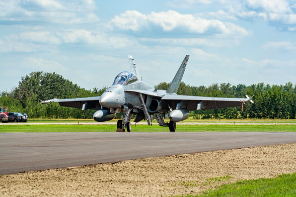 A military fighter in military grey paint parks and facing the photographer. There is green grass and green trees in the background. The sky is cloudy with some blue.