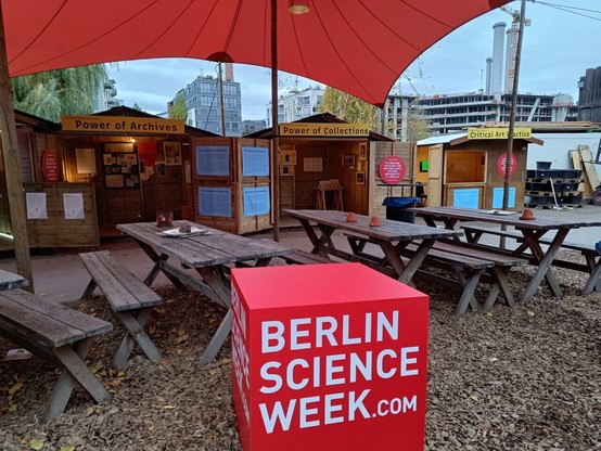 Bänke und Tische, davor ein großer Würfel in rot, auf dem Berlin Science Week Punkt com steht. Dahinter drei Markthütten auf denen drei Schilder sind:
"Power of Archives"
"Power of Collections"
"Critical Art Practice"

In den Hütten sind klein Poster an den Wänden zu erkennen.
