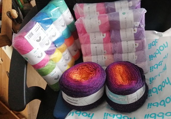Drei Wollpackungen, eine in Regenbogenfarben, die anderen beiden sind verschiedenen Töne von Pink und Lila. Außerdem zwei Bobbel in Farbverlauf von Violett zu Orange.