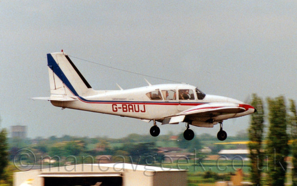 Side view of a cream and red, twin engined light aircraft flying from left to right at a very low altitude, with undercarriage lowered and nose down slightly, suggesting it is about to land, , with hangars, trees, and fields in the background, under a grey sky.