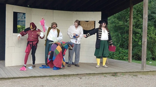 Five actors perform a commedia dell'arte play in front of a house scenic piece on a wooden outdoor stage during a medieval camping even portraying  the stock characters of Arlecchino (badly disguised as a fairy) sealing shoes from Pantalone (who is confused and annoyed) while Brighella and Oratio pretend to be amazed and il Dottore looks on with academic interest.