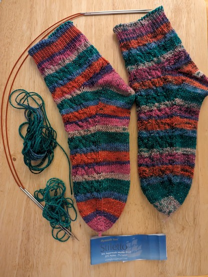 Two completed socks on their side showing the fleegle heel.  There are also two green bits of leftover yarn,  two chiaogoo red lace circular needles, and a label showing the yarn is stiletto by Plymouth