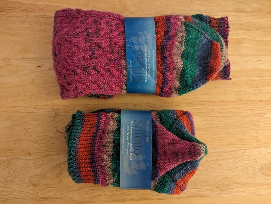 Two completed pairs of socks wrapped in the yarn bands that contained the yarn originally.  One is midcalf with the full width sole, and one is ankle length with the narrower sole.