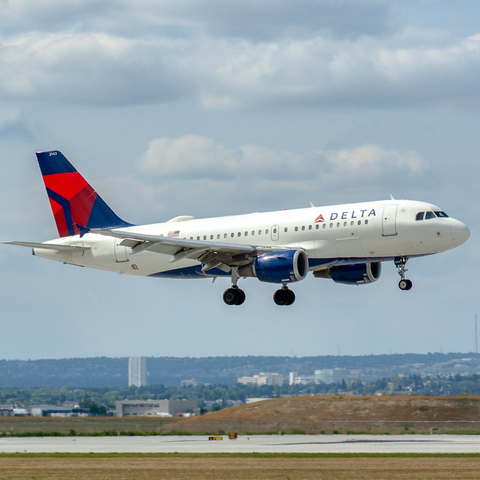 A white jetliner with blue belly and engines and a blue tail with the red delta logo. The sky is blue. The runway below the airplane. Airport buildings and the city in the background