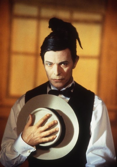 David Bowie wearing a a white shirt and a dark vest with watching bow tie and holding a hat over his chest with a black kitten on top of his head