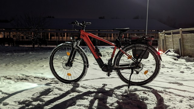 Ein rotes Raleigh Dundee Mountain Bike steht im Schnee auf einem Hof, es wird durch ein Flutlicht angestrahlt. Im Hintergrund Stallgebäude.