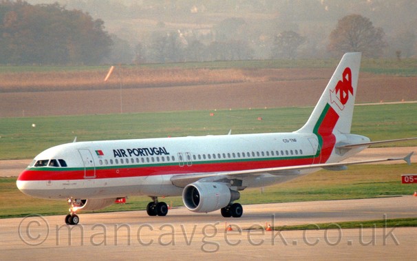 Side view of a white, red, and green twin engined jet airliner taxiing from right to left, in front of green fields leading up to trees in the background.