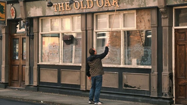 Main character, TJ, stands in front of his dilapidated pub, The Old Oak, a metaphor for the decrepit state of the UK under the Tories. He has is back to us and is try to straighten the letter 'K' in the golden letters that spell out the pub's name.