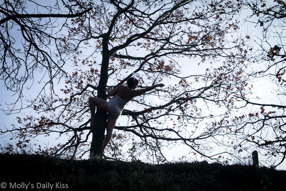 Molly wearing angel halo and white body stocking wrapped round a tree and pointing into the distance. The tree has a few autumn leaves left on it