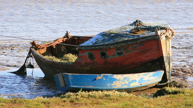 A disused boat and tender by the seashore. The boat is red above and black below, with three portholes. The tender, which lies in front of it, is blue. Both boats have peeling paint and look abandoned. There seems to be grass growing in the larger of the two.