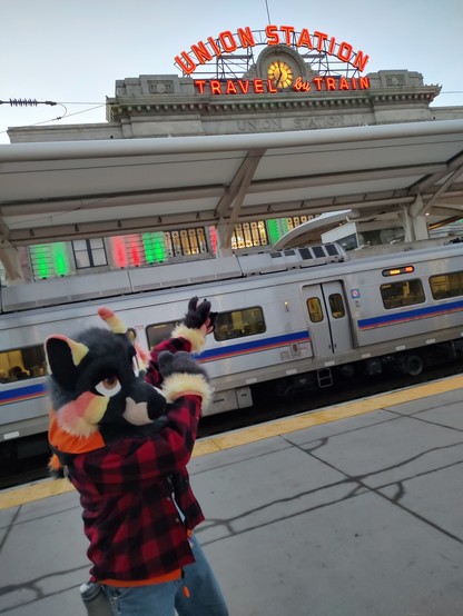 Coyote fursuiter with colorful ears, face, paws, and tail in red flannel poses pointing upward at the Union Station sign and clocktower at Denver's Union Station in downtown Denver. A commuter train is at the platform in the background.
