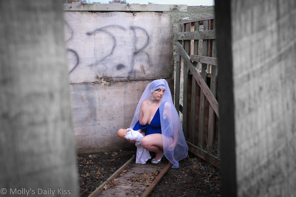 Looking through the side of a stable. Molly is crouching down in the corner holding a doll baby wrpped in white cloth. She is wearing a blue dress with the side pulled down exposing one breast, sparkly high heels and white headdress so she looks like the virgin mary