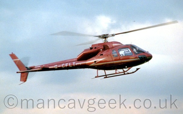 Side view of a brown helicopter with a light green and gold stripe running along the body, flying from left to right at a low altitude, in front of a background of blue skies and fluffy grey clouds.