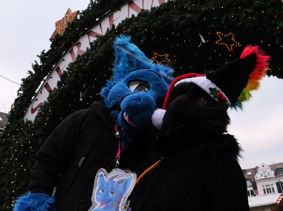 Me in my dragon fursuit partial and a muzzle harness, and MewAzunel in her partial fursuit, both looking to the camera, in front of a big semicircle of fir branches.