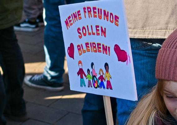 A child with long blonde hair looks down. Their face is only partially visible. They is holding a sign that says: "I want my friends to stay." Several people with different skin colours and a person in a wheelchair are painted underneath that phrase.