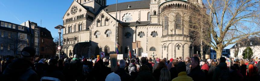 Panoramic photo of part of the demonstration. In the foreground many people with signs and flags, in the background the cathedral in Neuss. The sun is shining, the sky is blue.