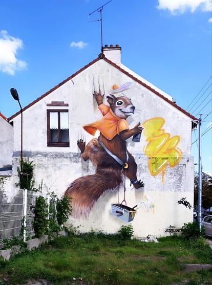 Streetartwall. A comic Style mural with a squirrel and with 3D effect was sprayed/painted on the outside wall of a white one-story house with a window on the left. The squirrel is wearing a yellow shirt and a baseball cap that flies above him. It hangs with its arms and legs clawed into the wall of the house. A square metal bucket hangs from a rope with a belt around its belly and dangles downwards. He looks around while holding a spray can with one hand and spraying wild yellow waves of graffiti on the wall. (There is a green front garden in front of the mural and a white garden fence with a lantern to the side).