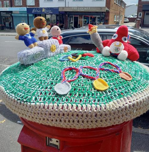 A knitted Postbox Topper to mark the Olympic Games. It has the olympic rings, medals, a winners' podium and a strange pink creature, carrying an Olympic torch, which I think represents the Paris 2024 mascot based on a Phrygian hat.