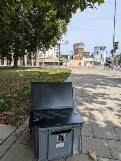 A box and a laptop on top. Electronics equipment in the box, some wires. Box is sitting on a sidewalk