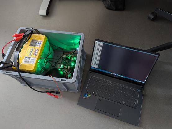 A photo of the elekctronics in a grey plastic box: A car battery, ZERO V2XNode and a laptop right next to it.