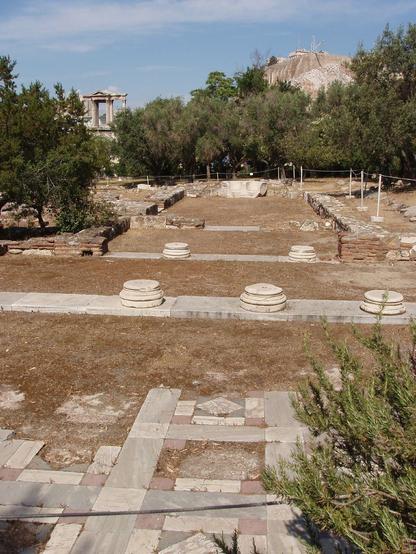 archaeological remains of bathing complex in Athens. Marble floor tiles and column bases, interspersed with trees. Beyond, Hadrian's Gate, and the acropolis.