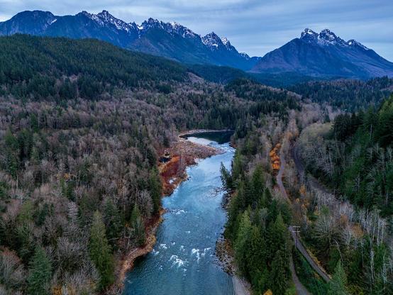 Color photo.  A river extends up from the bottom of the frame, turning a corner to the right at mid-frame.  On either side are bare trees, the hills above them dark with evergreens.  At the top of the frame is a line of snow-capped mountains.