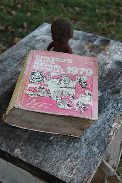 Outdoors atop a well worn wooden table sits a thick 1979 Chilton Auto Repair Manual and a very dirty vinyl doll.