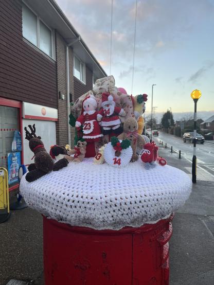 A British post box knitted topper showing various Christmas related knitted features, such as a reindeer, with advent numbers on.