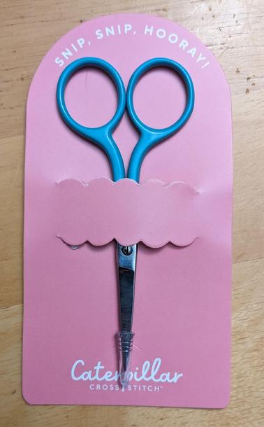 A pair of turquoise floss scissors in a pink Caterpillar Cross Stitch package.
