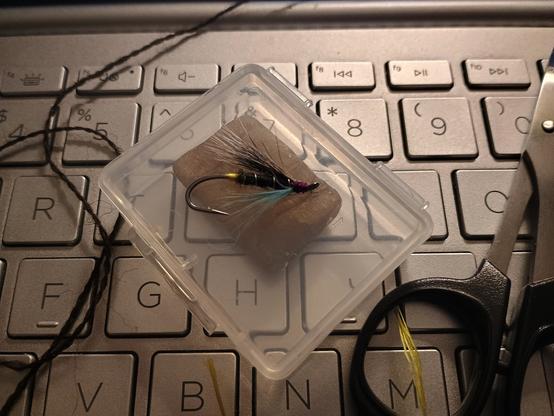 A fly fishing lure on a clear plastic box which is sitting on a keyboard. There's a piece of wool, a piece of yellow silk floss, and a pair of scissors also sitting on the keyboard.