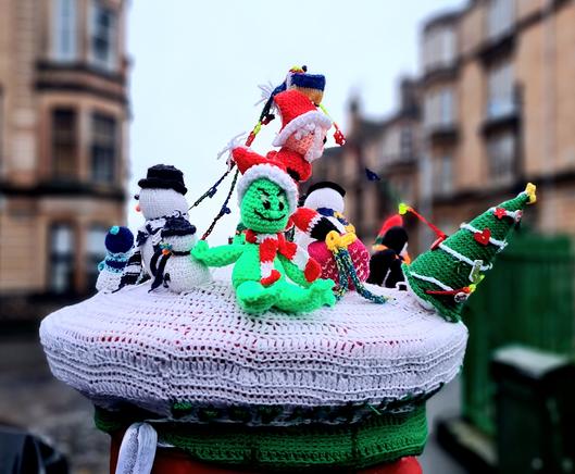 A knitted post box topper featurinh a grinch, a family of snowmen, some penguins, a Christmas tree and Santa Claus.