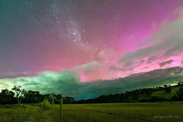 Some pink and faint partially-obscured-by-clouds beams from aurora australis on new years day, 2025. The foreground contains a field, wire fence and hills with trees.