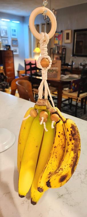 Macrame banana hanger, using a white cotton rope attached to a pale wooden ring. There is a darker wooden bead in the middle of the knot design and at the end of each cord is attached another dark wooden bead. All the beaded cords together support the weight of 2 banana bunches hung by their crowns.